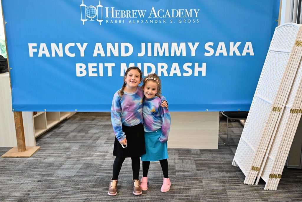 Two kids hugging happily in front of the Hebrew Academy backdrop banner at the opening event.