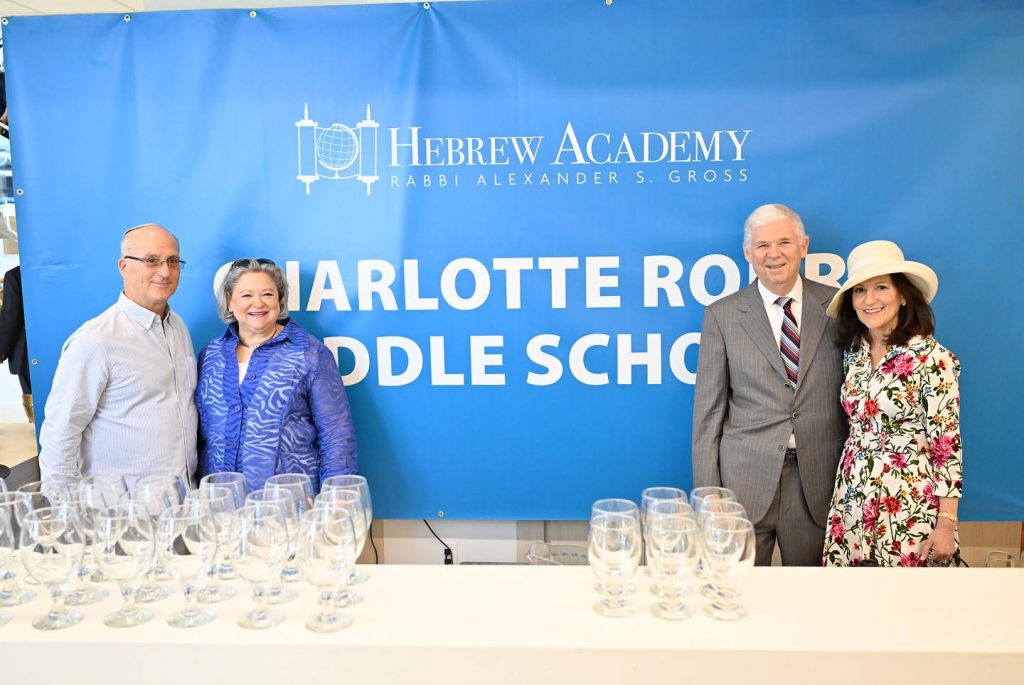 Two smiling couples standing in front of a Hebrew Academy backdrop.