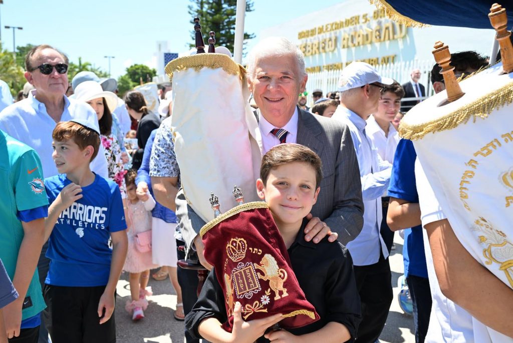 Adult and child proudly holding a Torah scroll at an outdoor event.