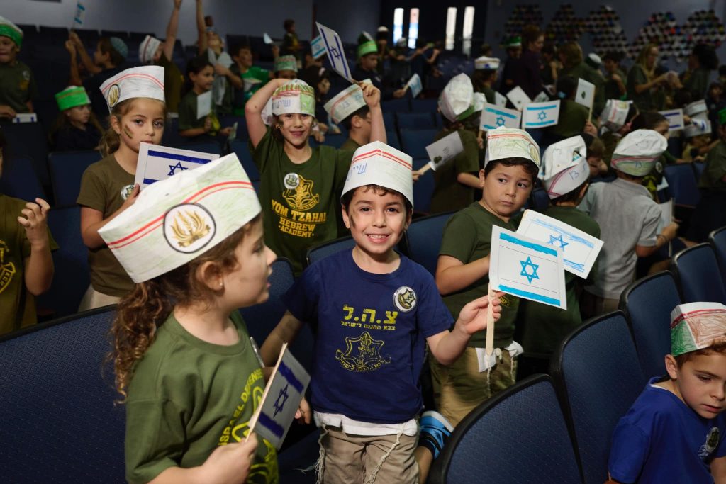 Group of kids indoors happily smiling and waving small Israeli flags while wearing paper hats.