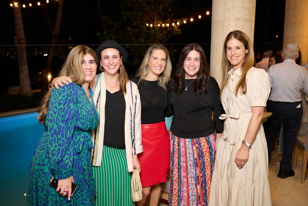 Five women happily posing for a photo at an outdoor evening event.