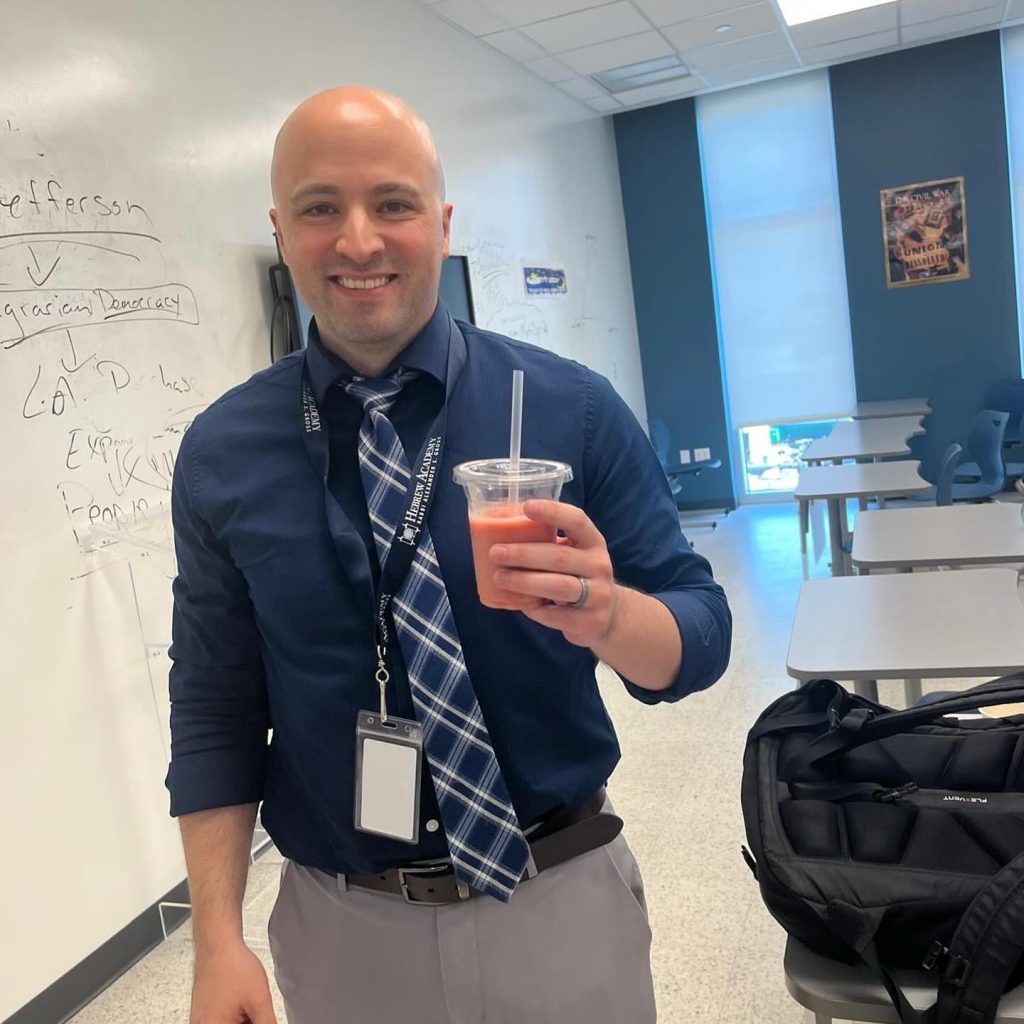 Male teacher smiling and holding a drink in a classroom during Teacher Appreciation Week.