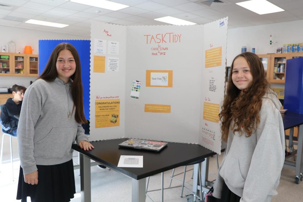 Two students in a classroom happily standing in front of their presentation.